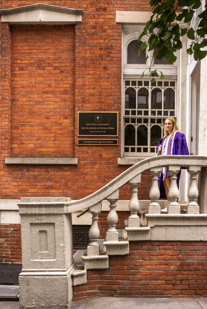 A student dressed in the NYU graduation gown walks down the steps of the NYU Silver School of Social Work building in New York City.